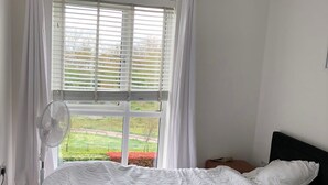 A bedroom with a bed, white curtains, a window with blinds, a standing fan, and a view of a garden.