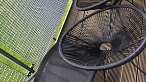 A rooftop terrace with a metal grill, a black mesh chair, and a view of a green lawn and trees.