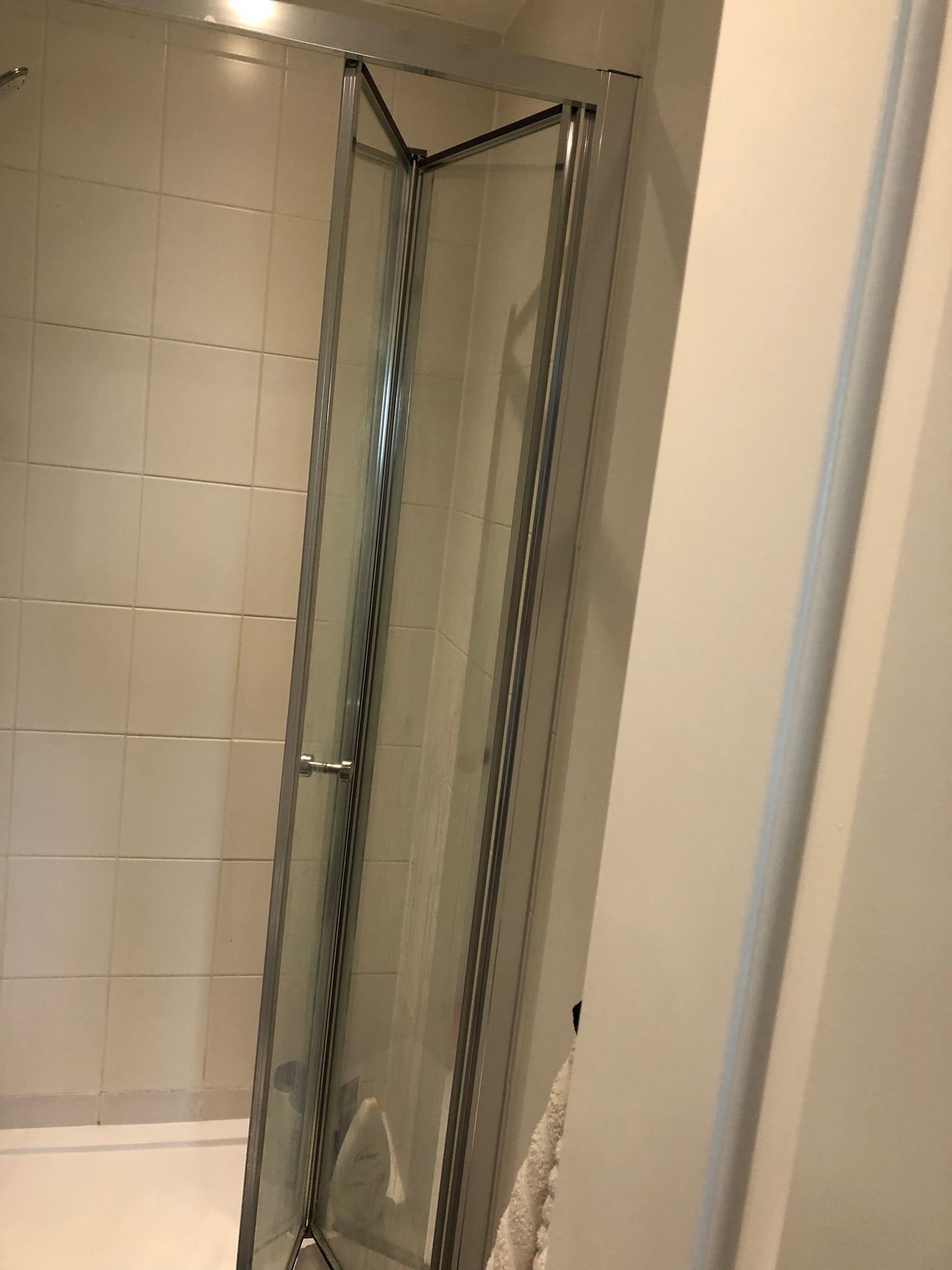 A glass-enclosed shower stall with a tiled wall and a white bathtub in the background.