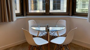 A dining area with a glass table and four white chairs, large windows with curtains, and a view of modern buildings.