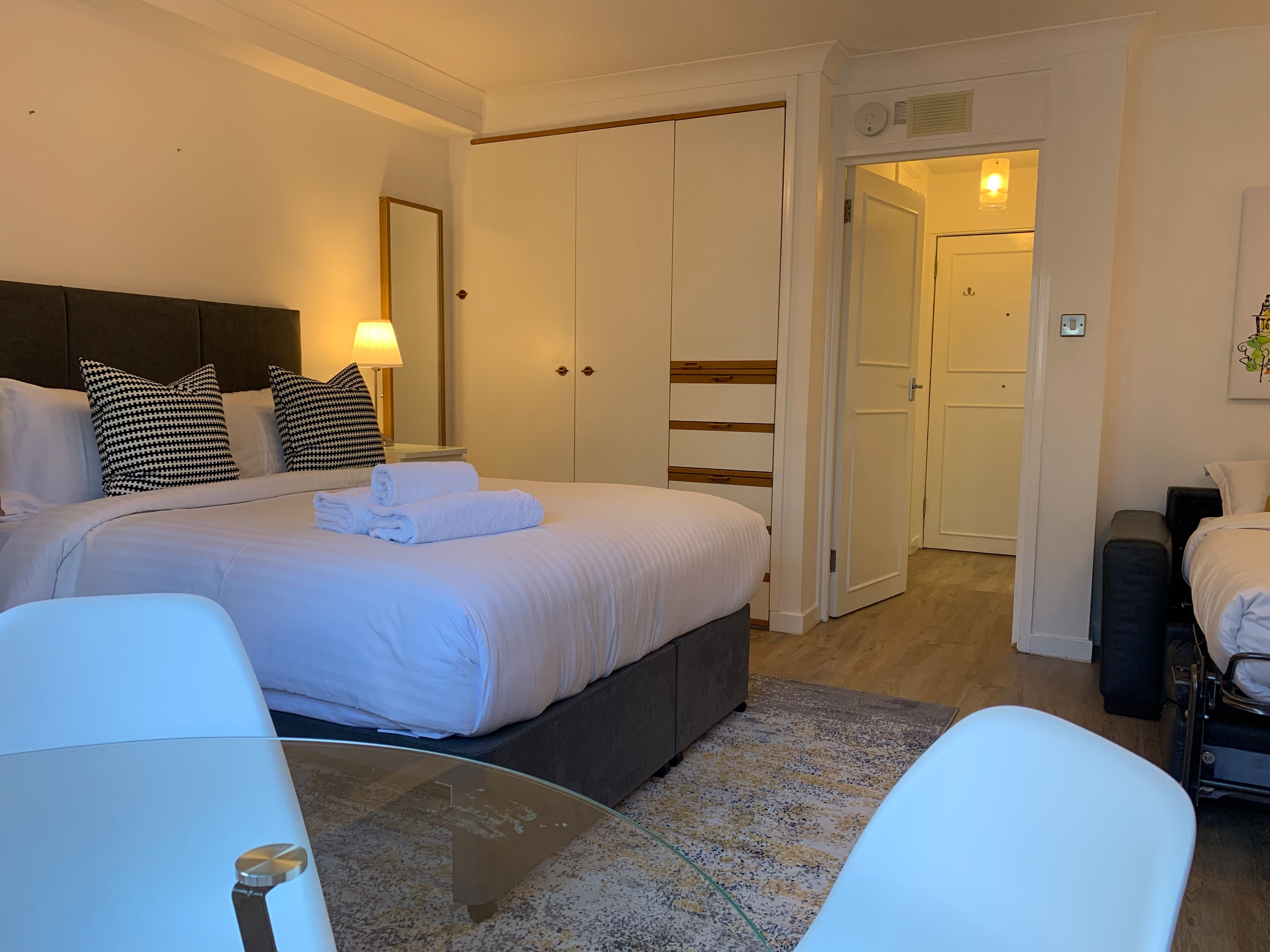 Una habitación de hotel con cama, mesita de noche, lámpara y una puerta que da a otra habitación.