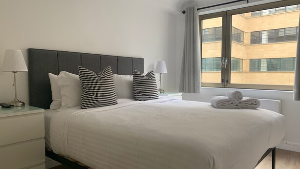 A hotel room with a bed, two bedside lamps, a window with curtains, and a view of a building outside.