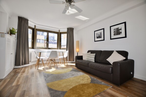 A modern living room with a dark brown sofa, a dining table with chairs, and a large window with curtains.