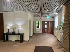A hallway with a wooden floor, a desk with a lamp and flowers, a door with a mirror, and a plant.