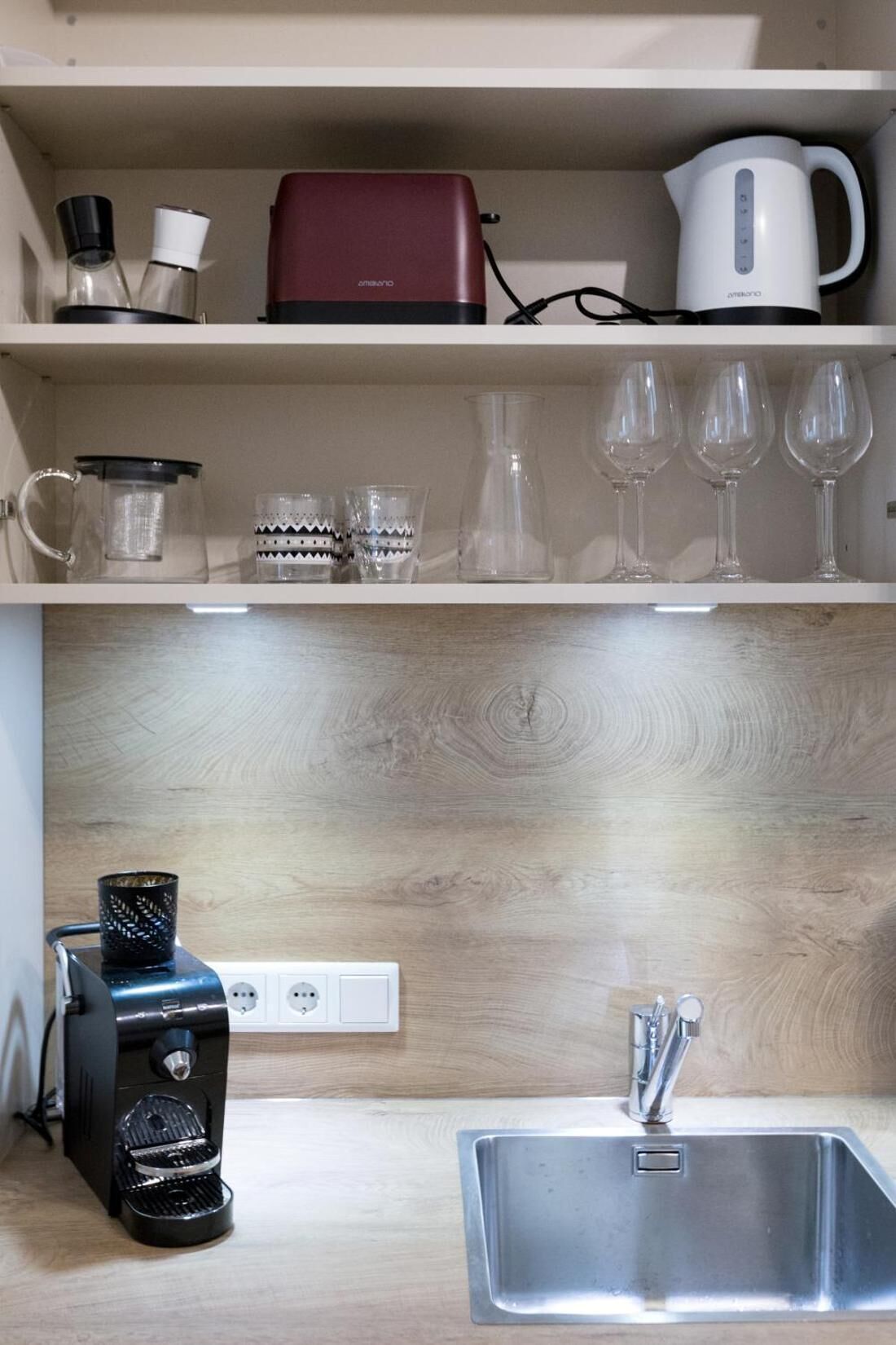 A kitchen with a coffee maker, a kettle, and wine glasses on the shelves.