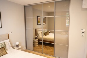 A bedroom with a large sliding glass door closet, a bed with white bedding, a bedside table with a lamp, and a small round table with a lamp. - Modern stylish first floor flat in Fitzrovia (Greater London)
