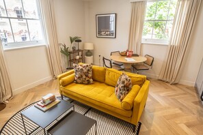 Um sofá amarelo com almofadas estampadas, uma mesa redonda pequena com cadeiras, um abajur e um quadro na parede.