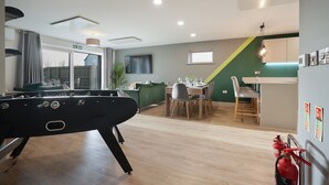 A modern living room with a foosball table, a dining area with a table and chairs, and a kitchen area with a window and a green accent wall. - High spec, 4 bed family eco-home w/private gym & tennis courts in Lincolnshire. (Lincoln)