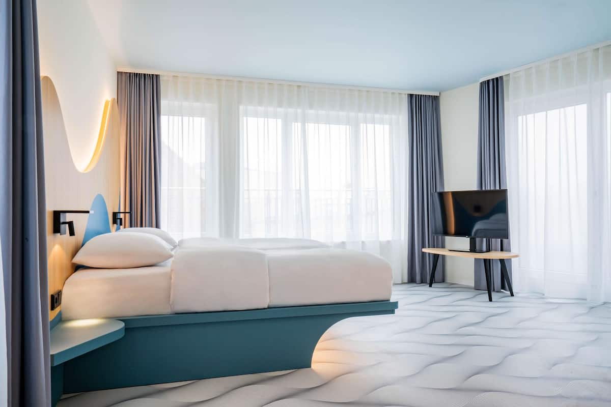 Une chambre d’hôtel moderne, équipée d’un lit, d’un téléviseur posé sur un meuble et de grandes fenêtres avec des rideaux.