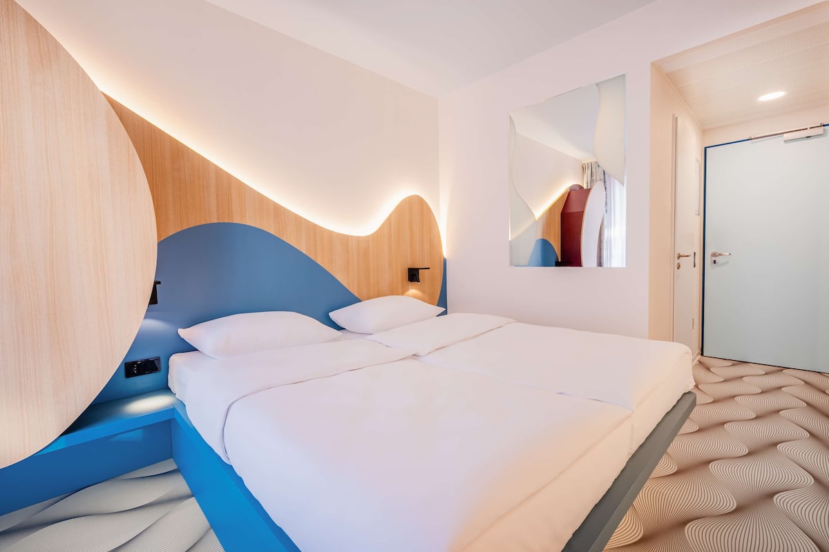 Une chambre d’hôtel moderne avec un grand lit, un mur décoratif en bois, un miroir et une moquette à motifs.