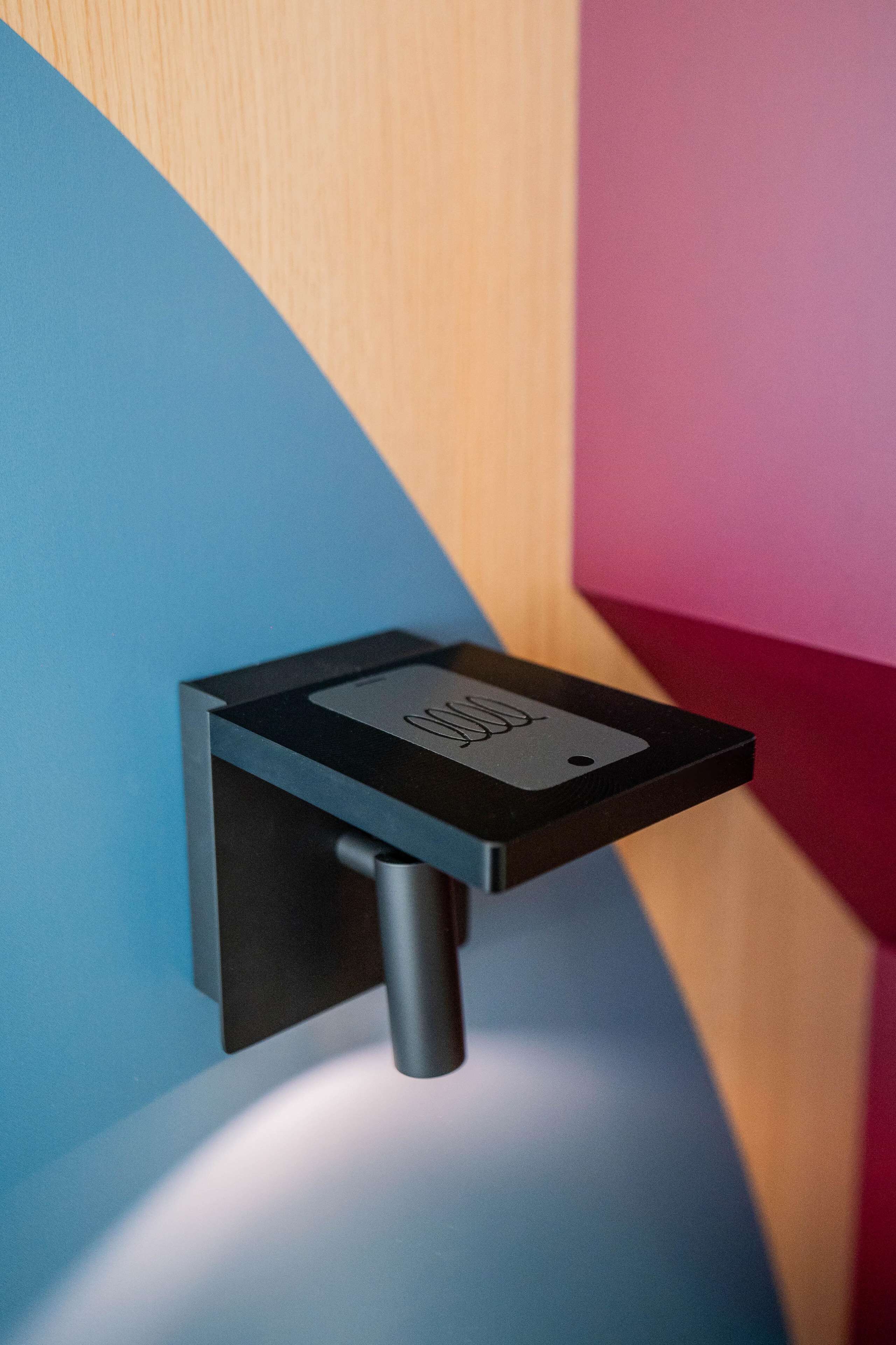 A wall-mounted device with a black base and a rectangular panel, possibly a control panel or sensor, against a wall with a blue and pink color scheme.