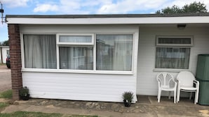 A single-story building with a white exterior, two windows with curtains, a small porch area with two white plastic chairs, and a green trash can. - 2-bed Seaside Retreat - Sleeps 5 & Parking (Great Yarmouth)