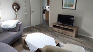 A living room with a sofa, a TV on a wooden stand, a white table, and a door leading to a kitchen.