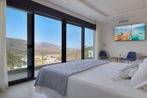 Une chambre avec un grand lit, deux fauteuils, un téléviseur et une vue sur un paysage côtier.