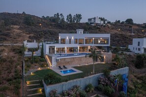 Une maison moderne à deux étages, dotée d’une piscine, entourée d’un jardin aménagé et d’un paysage vallonné.