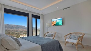 Een moderne slaapkamer met een groot bed, twee rieten stoelen, een televisie aan de muur en uitzicht op bergen door grote ramen.