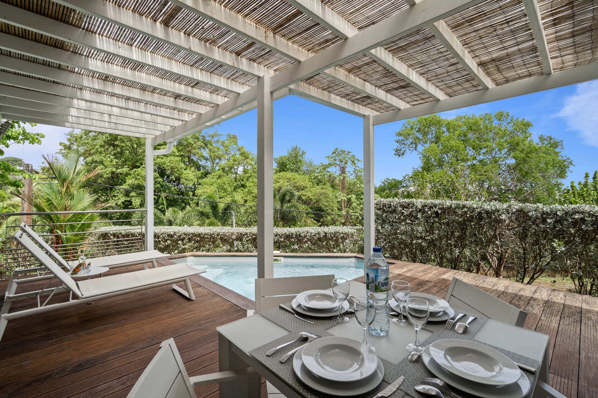 An outdoor dining area with a pool, surrounded by greenery and a pergola.