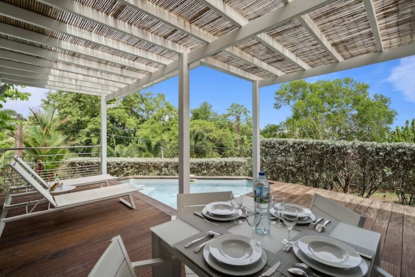 An outdoor dining area with a pool, surrounded by greenery and a pergola.