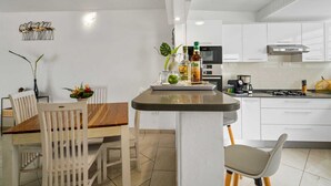 Una cocina moderna con armarios blancos, una isla central con un lavamanos, estufa y cafetera. Hay un comedor con una mesa de madera y sillas blancas.