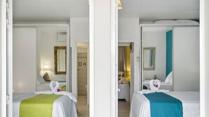 Habitación de hotel con dos camas, cada una con cubrecama blanco y toalla decorativa con forma de cisne. La habitación tiene un espejo, una mesita de noche y una puerta que da a otra habitación.