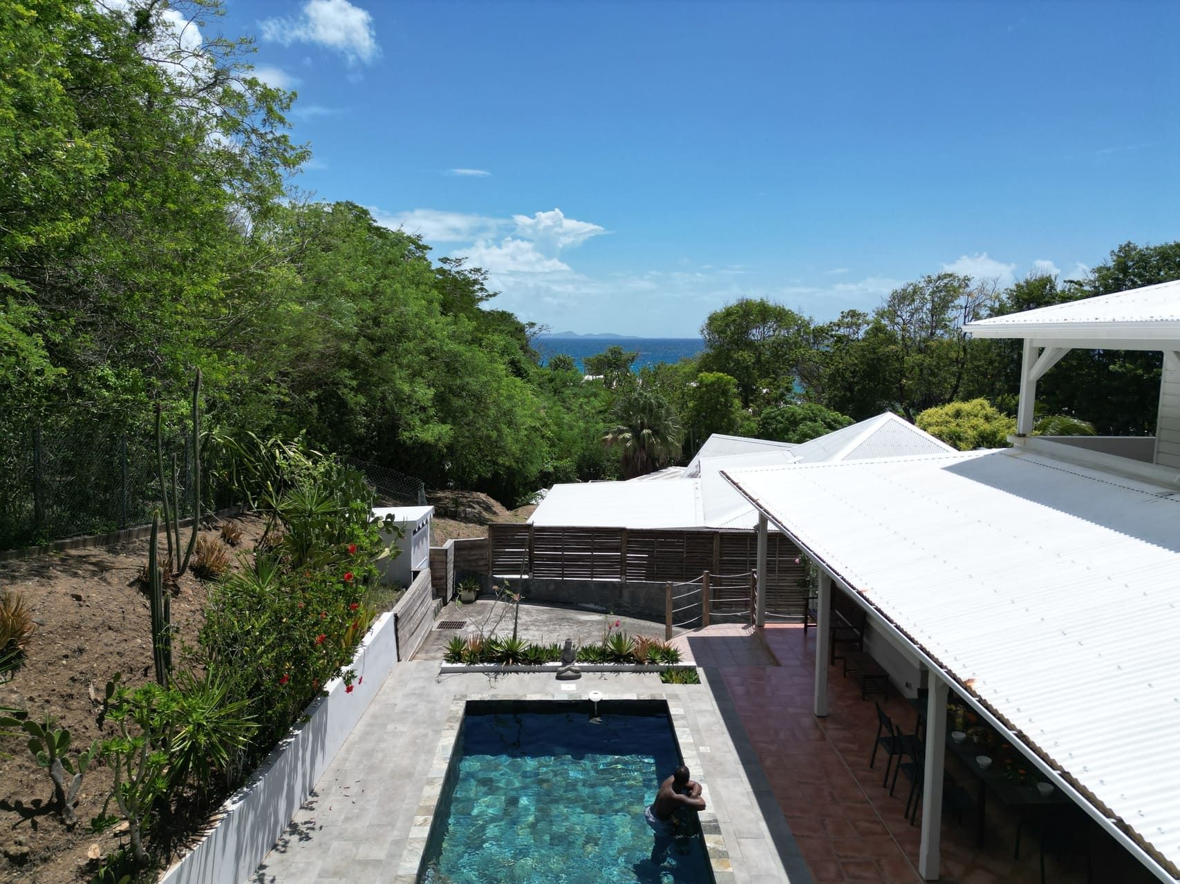 Un espace piscine offrant une vue sur l’océan, entouré d’une végétation luxuriante et d’un bâtiment blanc doté d’un patio couvert.