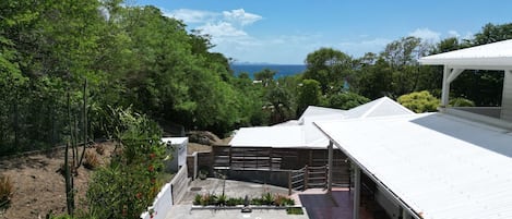Poolområde med udsigt over havet, omgivet af frodig vegetation og en hvid bygning med en overdækket terrasse.