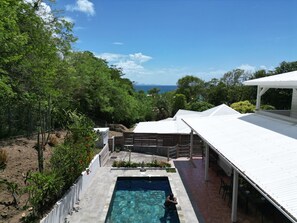 Un área de piscina con vista al océano, rodeada de exuberante vegetación y un edificio blanco con un porche cubierto.