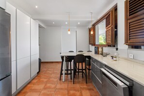Una cocina moderna con armarios blancos, un refrigerador de acero inoxidable, una mesa de comedor con sillas, luces colgantes y alacenas de madera sobre la encimera.