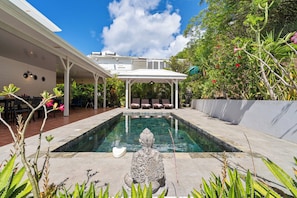 A swimming pool with a stone statue in the center, surrounded by lush greenery and a covered seating area.