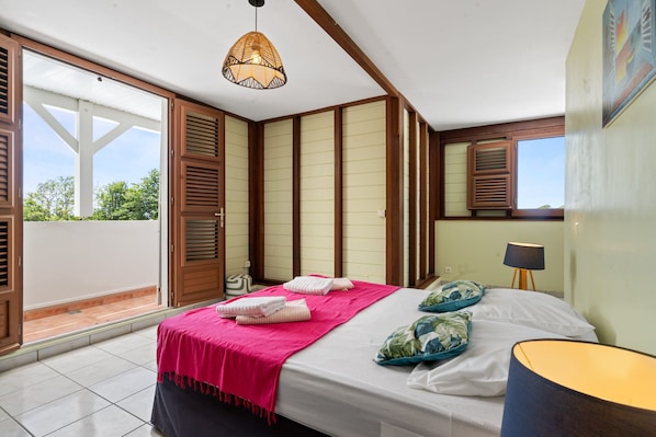 A bedroom with a bed, a bedside lamp, a window with shutters, and a view of the outdoors.