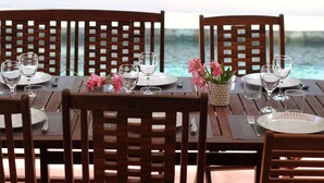 Una mesa de comedor preparada para cuatro personas, con sillas de madera, un jarrón de flores rosadas y vista al océano.
