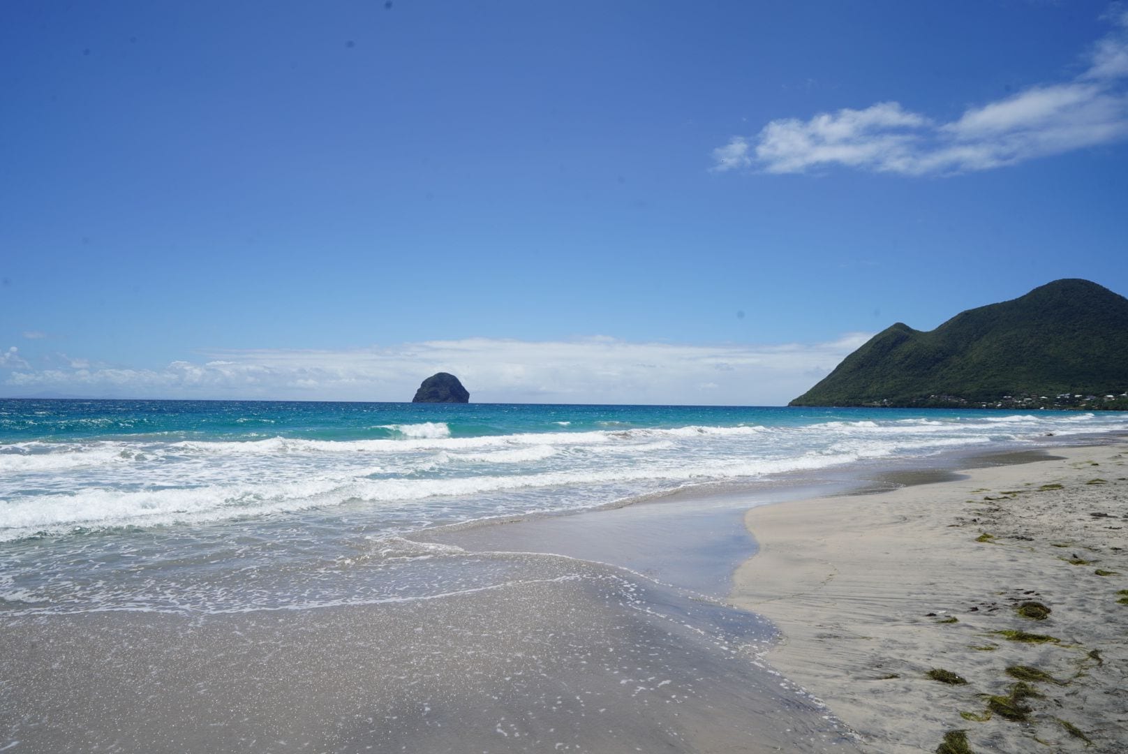 A beach with clear blue water, a sandy shore, and a mountain in the background.