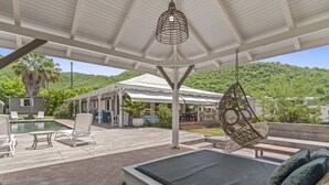A covered outdoor area with a pool, lounge chairs, a hanging chair, and a bed with pillows.