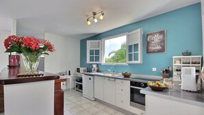 A modern kitchen with white cabinets, a teal accent wall, a coffee-themed sign, and a window with white shutters. - Villa Kaline - Three Bedroom Villa, Sleeps 6 (Le Vauclin)