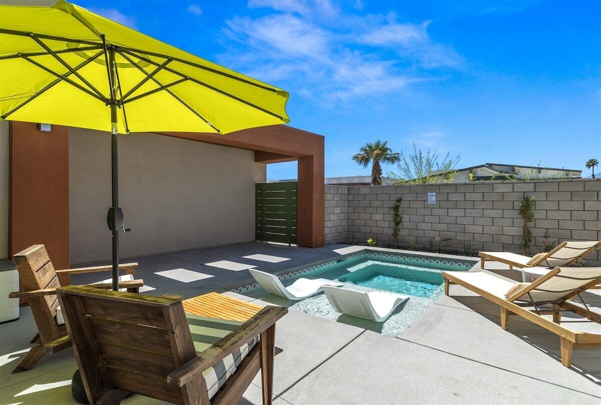Zona de piscina amb gandules de fusta, un parasol groc i una petita banyera d'hidromassatge.