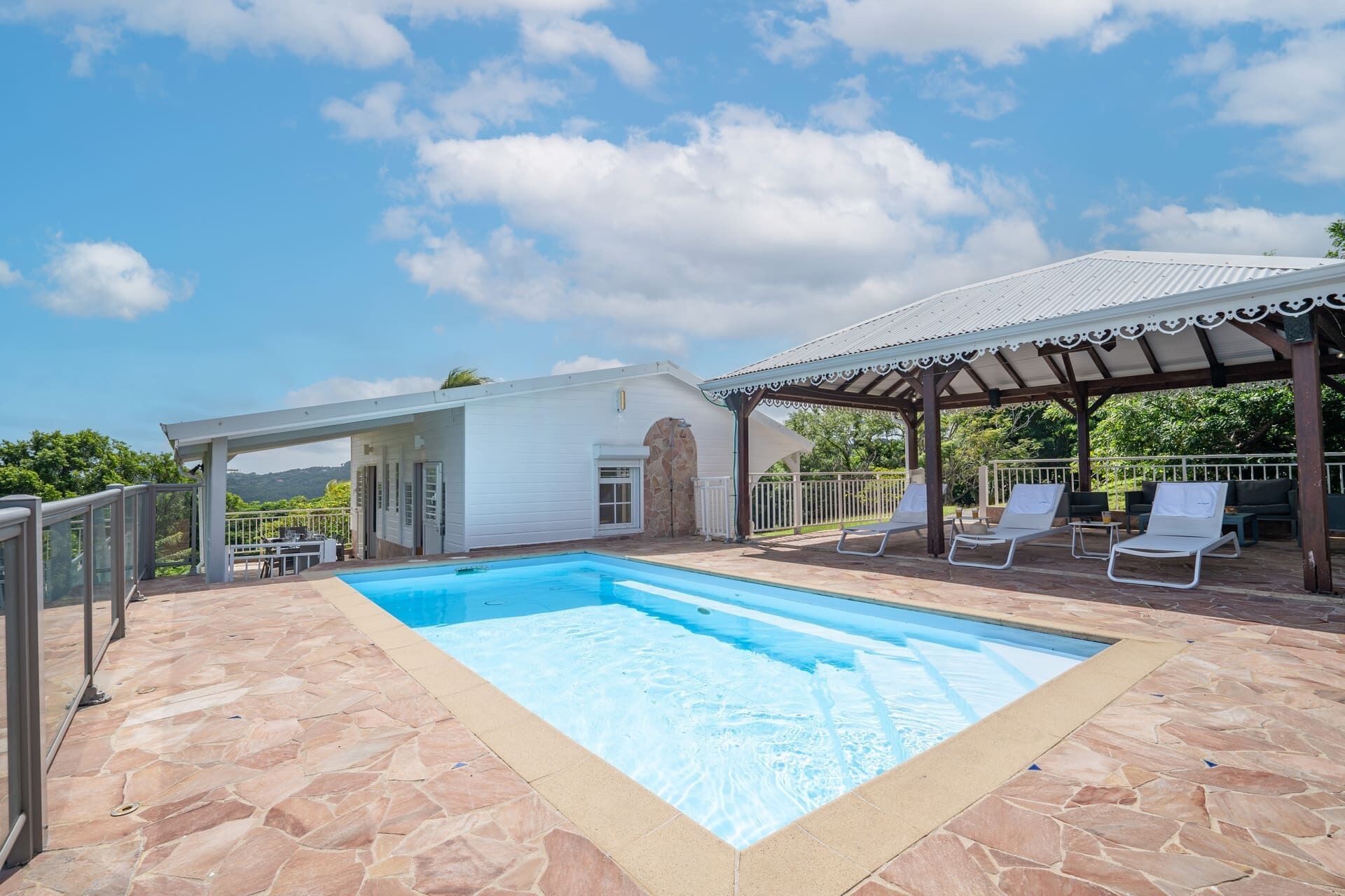 A swimming pool with stone tiles, a covered patio with lounge chairs, and a white building with a stone archway.