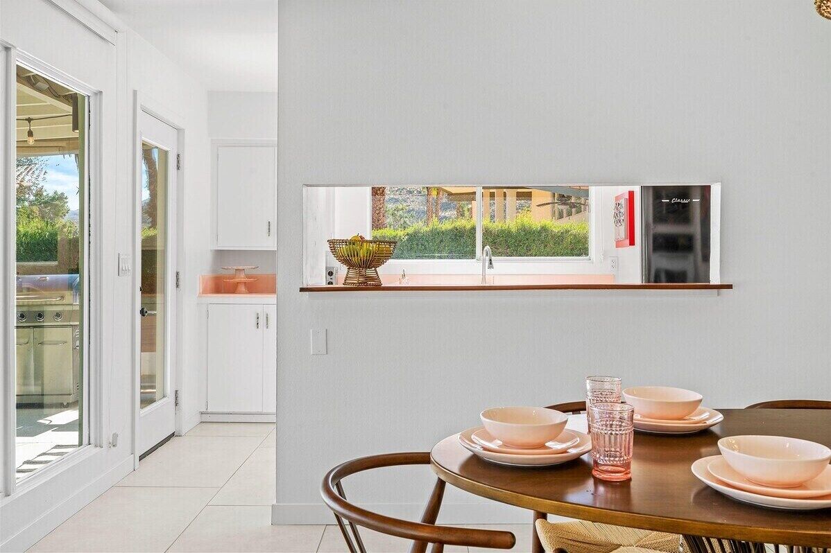 A modern kitchen with a built-in refrigerator, a dining table with pink plates, and a view of greenery outside.