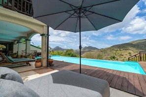 A poolside area with a wooden deck, lounge chairs, a large umbrella, and a view of mountains.
