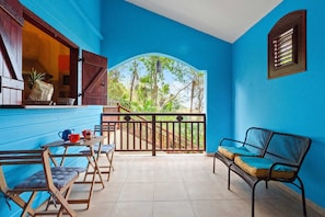 A room with a blue wall, a balcony with a view of trees, a table with chairs, and a bench.