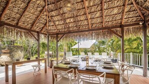 Un área de comedor al aire libre con techo de paja, sillas de plástico blanco y una mesa preparada para una comida.