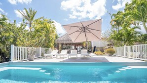 Un área de piscina con sillones de descanso, una estructura de techo de paja y un paraguas grande.