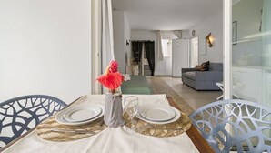 Ein Essbereich mit einem für zwei gedeckten Tisch, einer Vase mit roten Blumen und Blick in ein Wohnzimmer mit einer Couch und einem Wandkunstwerk.