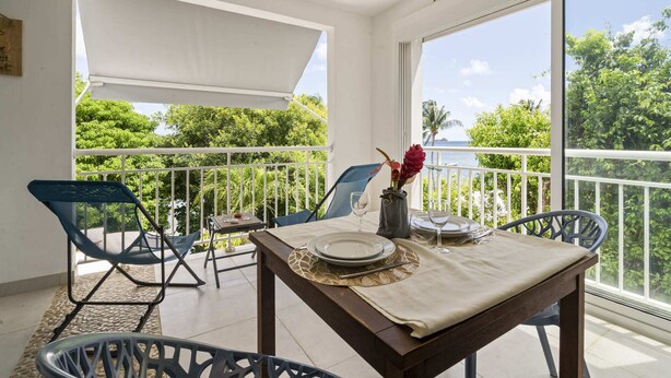 Ein Balkon mit Tisch für zwei Personen, Stühlen, einem kleinen Tisch mit Blumenvase und Blick auf Meer und Grünflächen.