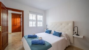 A hotel room with a large bed, a wooden door, a window with bars, and a bedside table with a lamp. - Casa Monash (Puerto del Carmen)