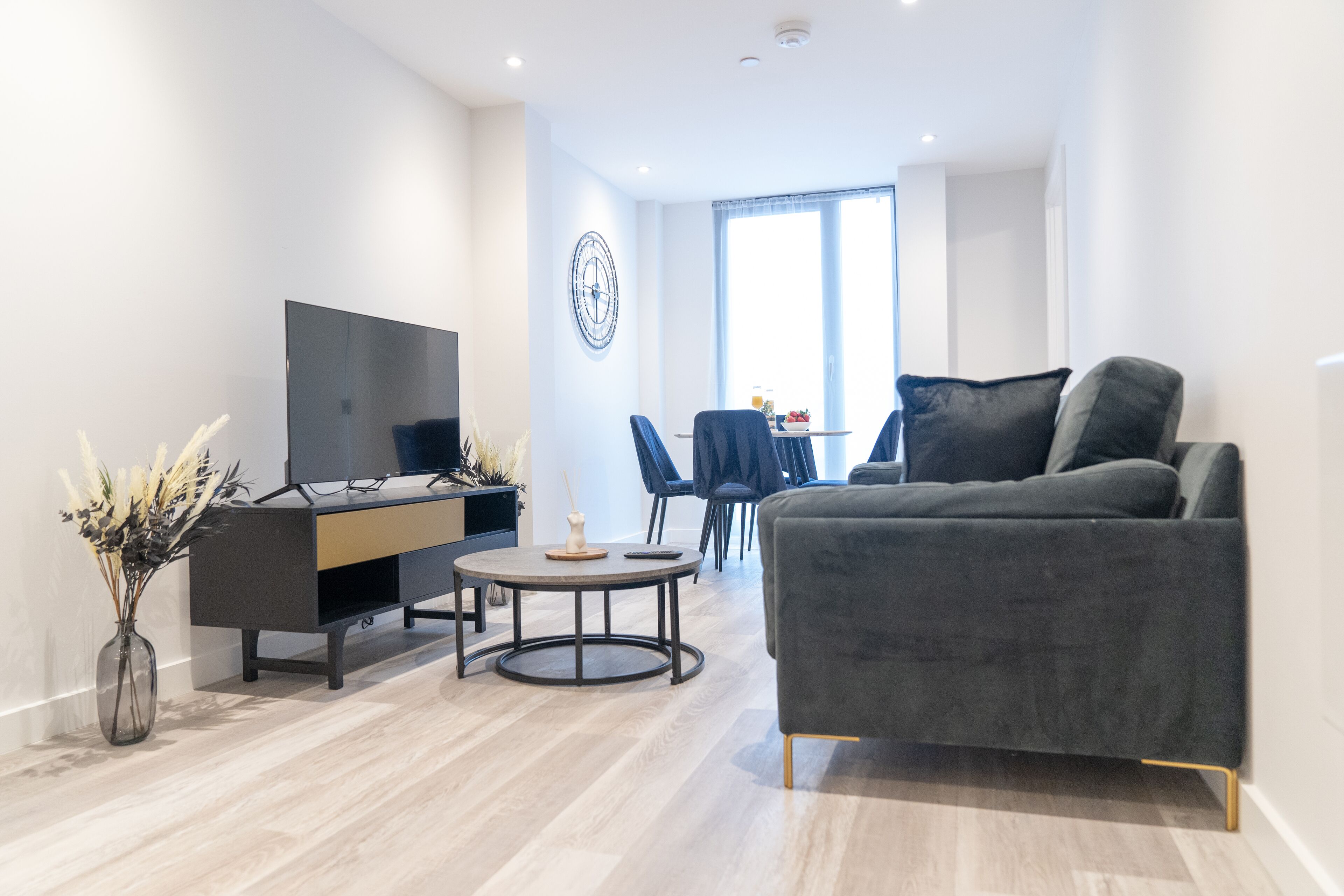 En moderne stue med en grå sofa, et rundt sofabord, et fjernsyn på et sort stativ og et spiseområde med stole og bord.