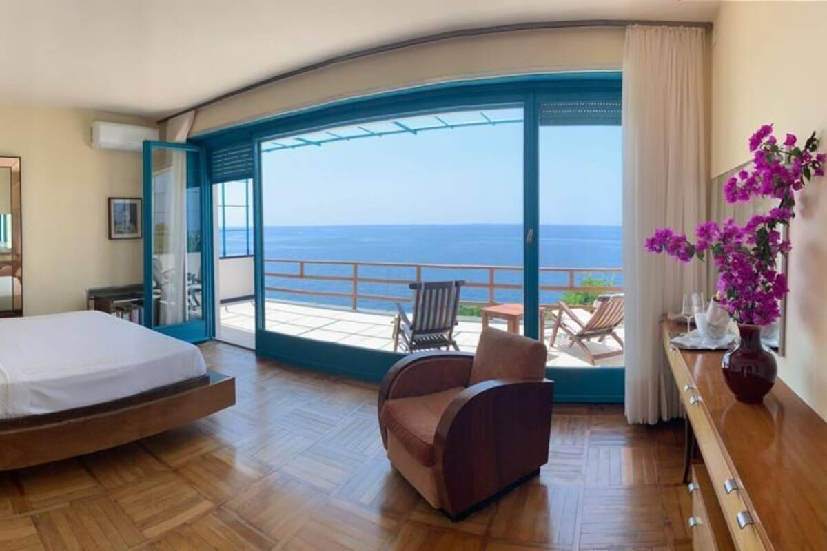 A bedroom with a bed, a chair, a desk, and a view of the sea.