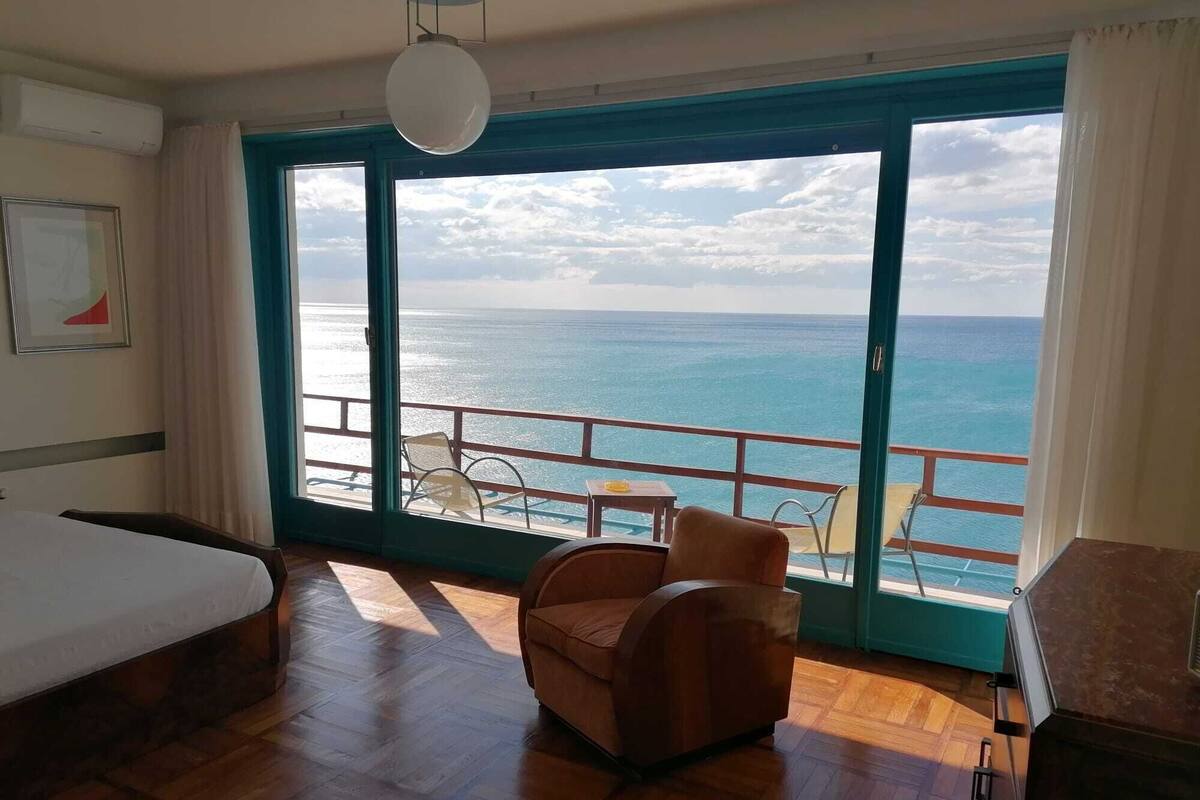 A room with a large sliding glass door leading to a balcony with a view of the ocean. There is a bed, a chair, and a small table on the balcony.
