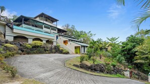 Un edificio de dos pisos con un camino de entrada curvo, rodeado de vegetación tropical y rocas.