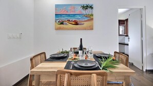 Un coin repas avec une table en bois dressée pour quatre personnes, entourée de chaises en osier. Un tableau représentant une scène de plage est accroché au mur au-dessus de la table.