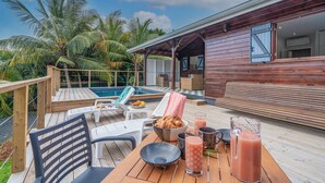 Une terrasse en bois avec une piscine, des chaises longues et une table où sont disposés des aliments pour le petit-déjeuner.